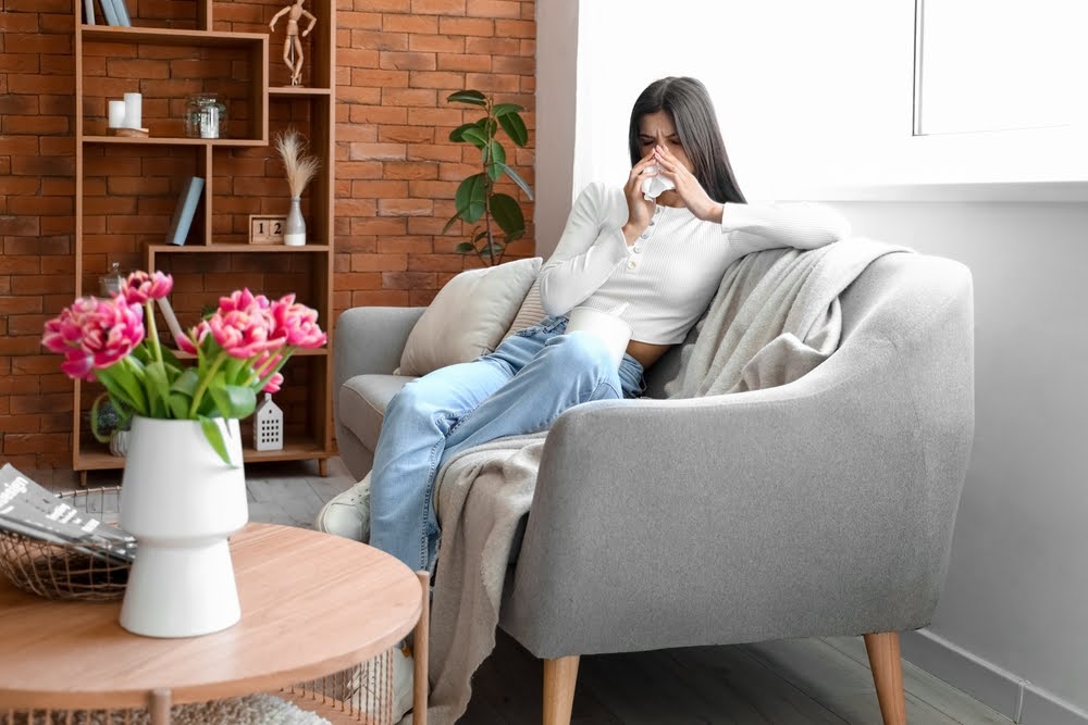 A young woman suffering from allergies and blowing her nose inside her house on the couch