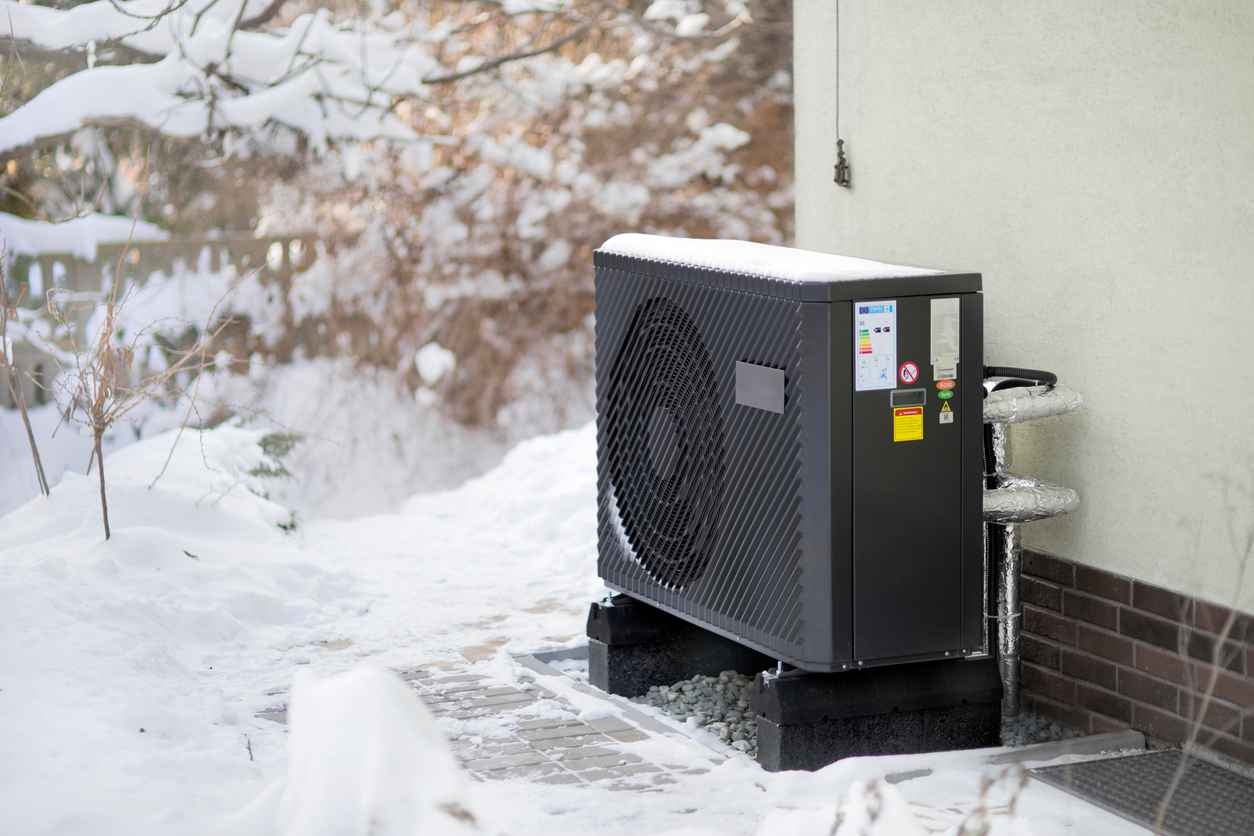 Photo of a modern air source heat pump unit standing against the residential home. Shot in winter, in a snowy setting.